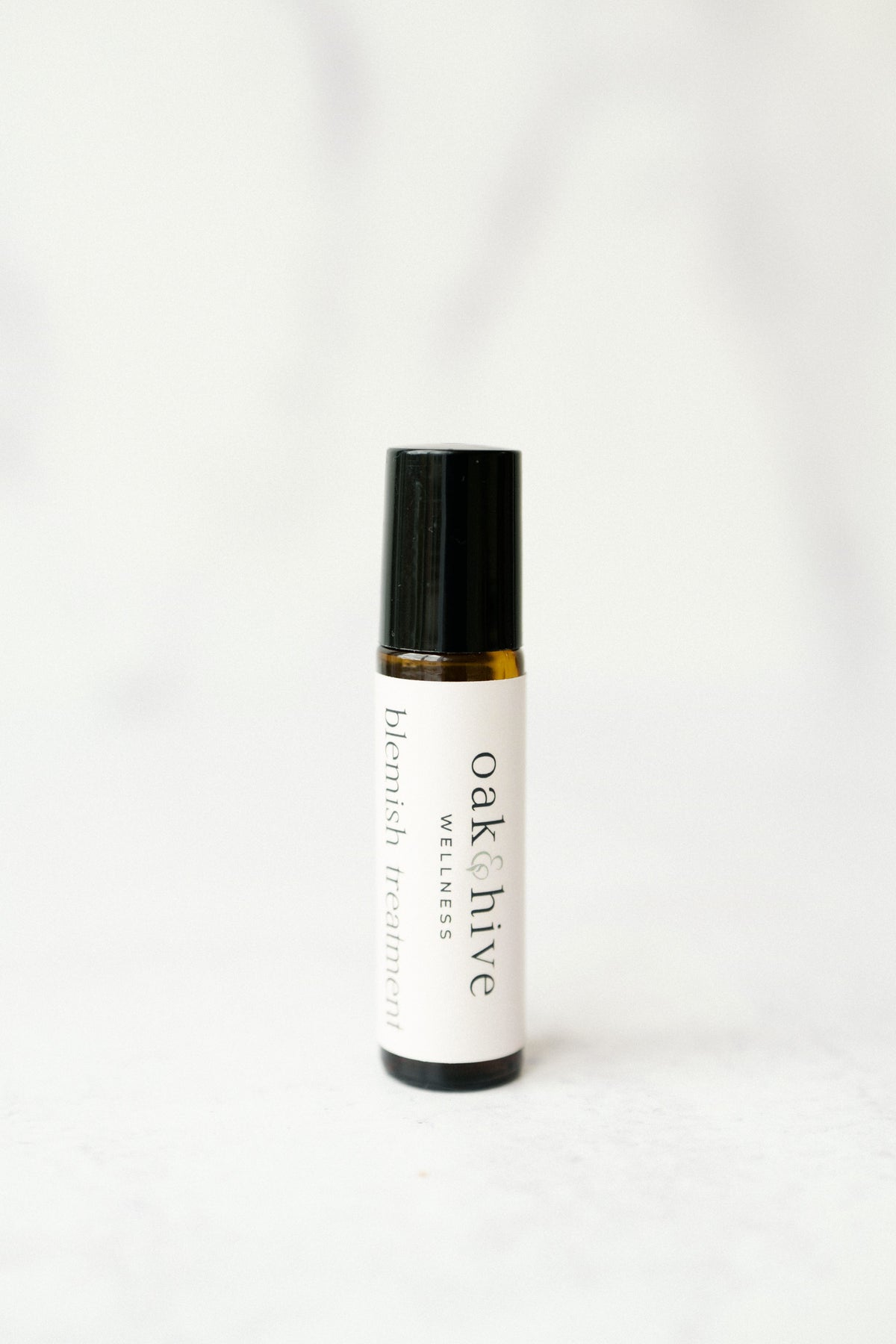 A amber colored roller bottle with a white label and gray letters reading 'Oak & Hive Wellness Botanical Blemish' on a white table and white background.