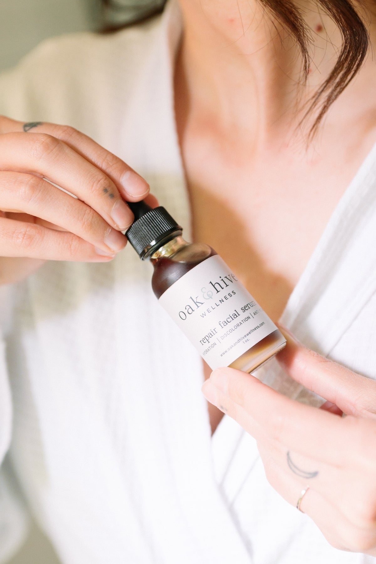 Person holding a bottle of skincare serum in a white robe with a white label.