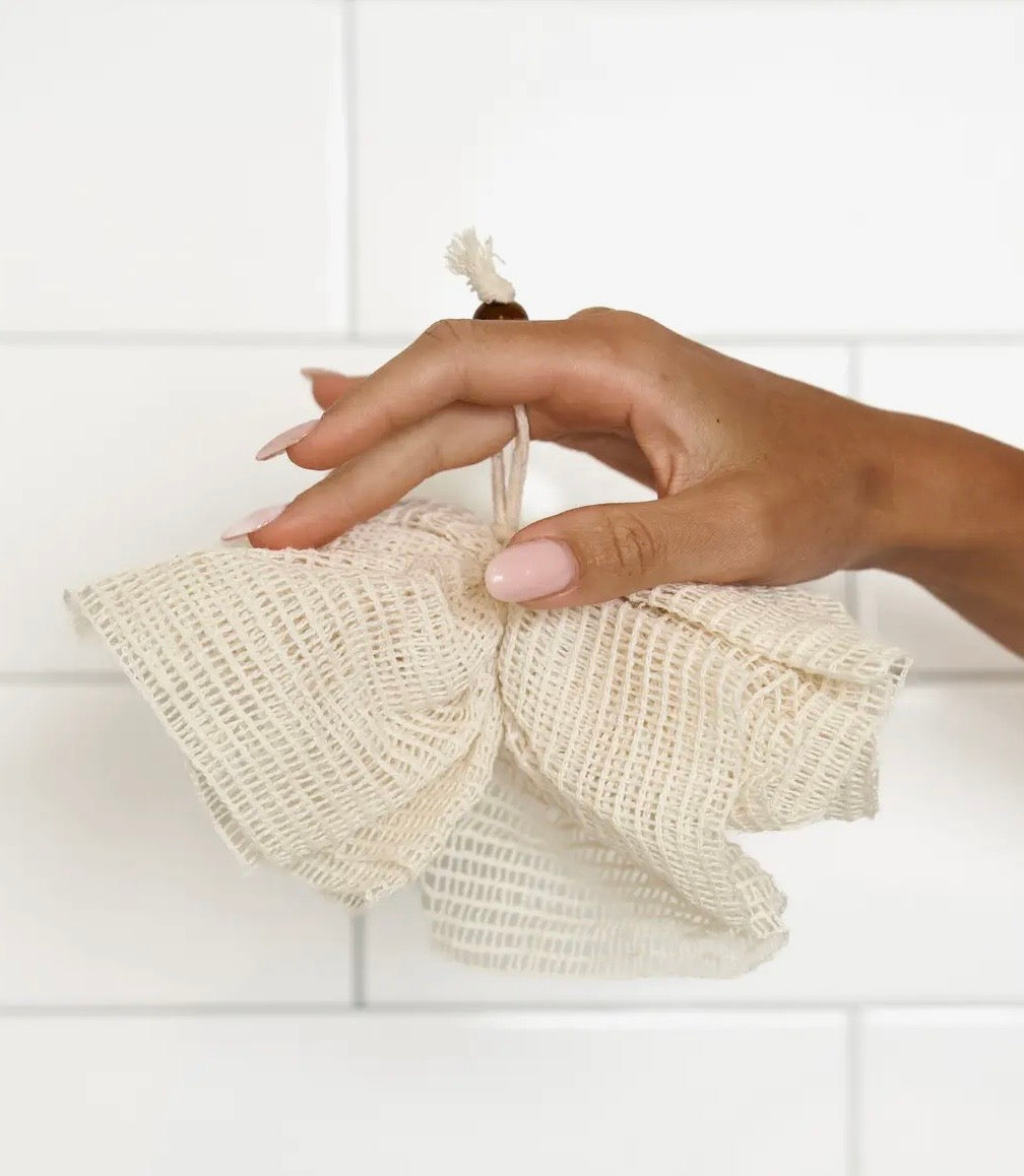 Hand holding a beige reusable sisal shower pouf against a white tiled wall.