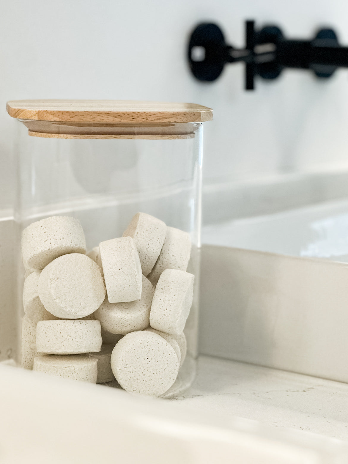 Clear glass jar with wooden lid filled with white tablets on a light surface.