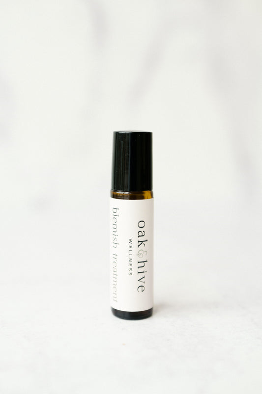 A amber colored roller bottle with a white label and gray letters reading 'Oak & Hive Wellness Botanical Blemish' on a white table and white background. 