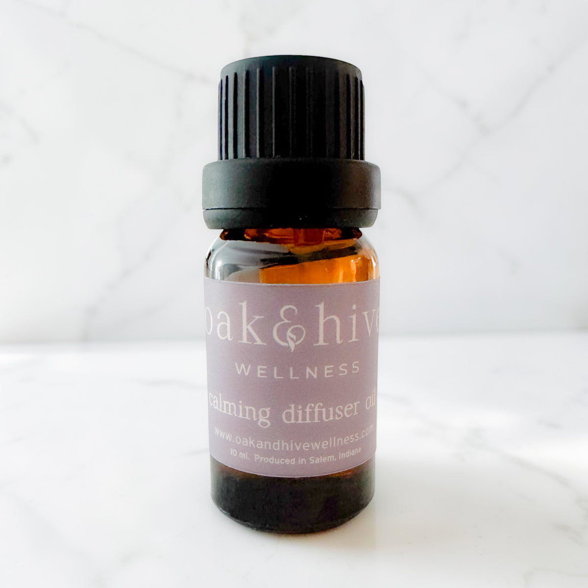 A small amber glass bottle with a black ribbed cap and a light purple label reading 'oak & hive wellness' and 'calming diffuser oil,' photographed on a light marble-like surface and gray background.