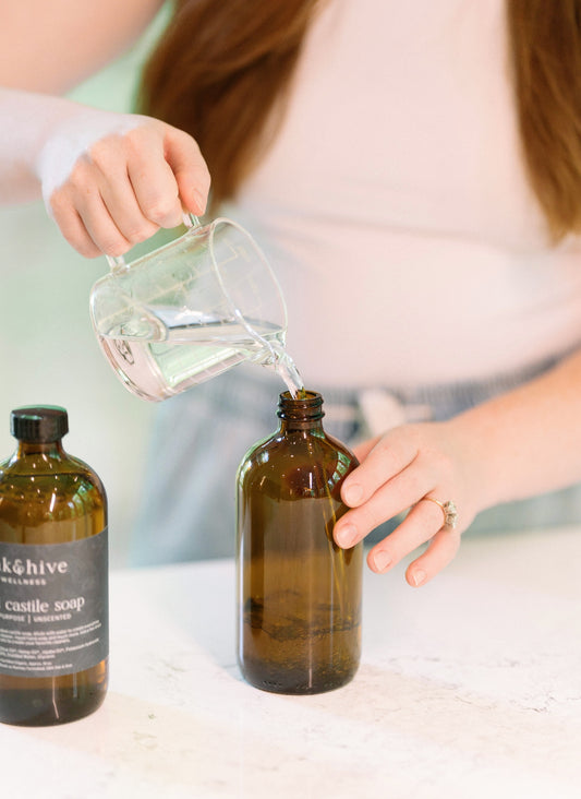 Person pouring liquid from a glass container into an amber bottle with a visible brand label.