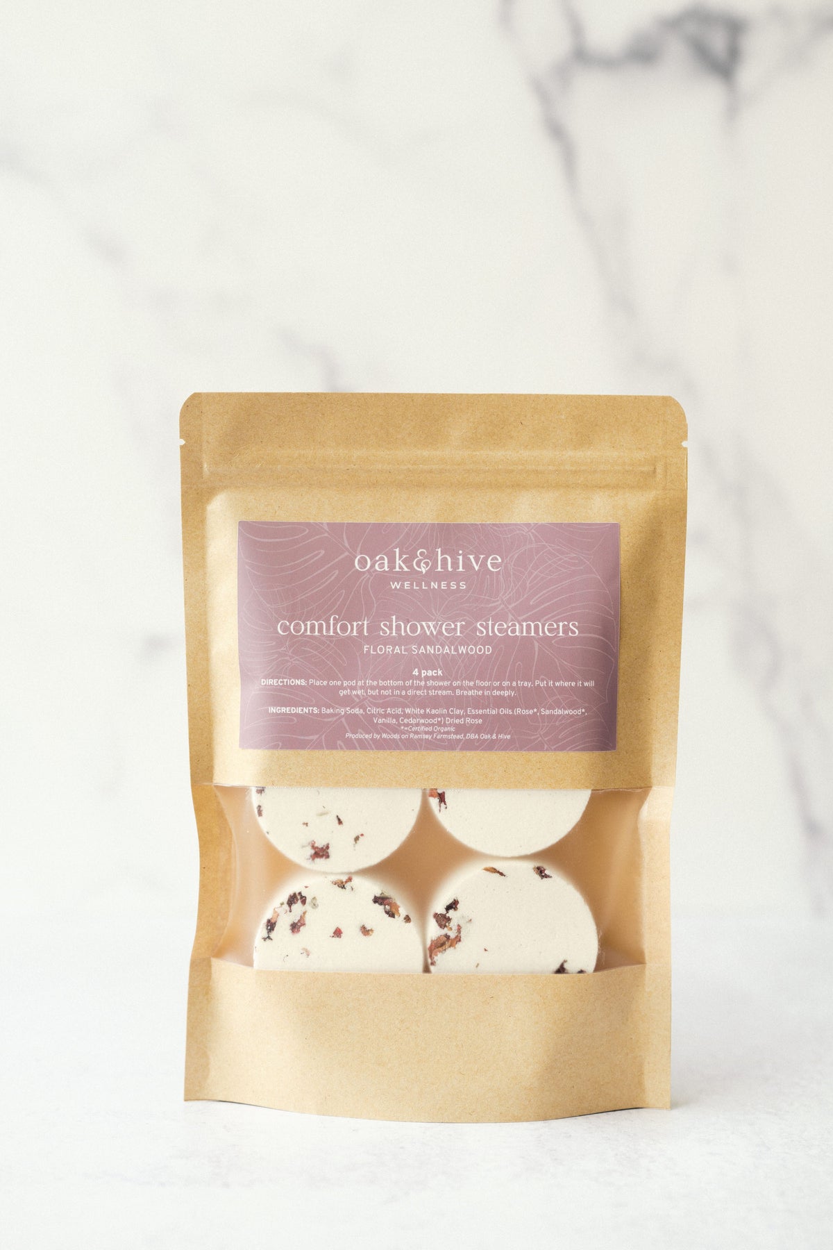 a brown package with white pucks with dried rose petals on them with a mauve pink label on a white marble background. 