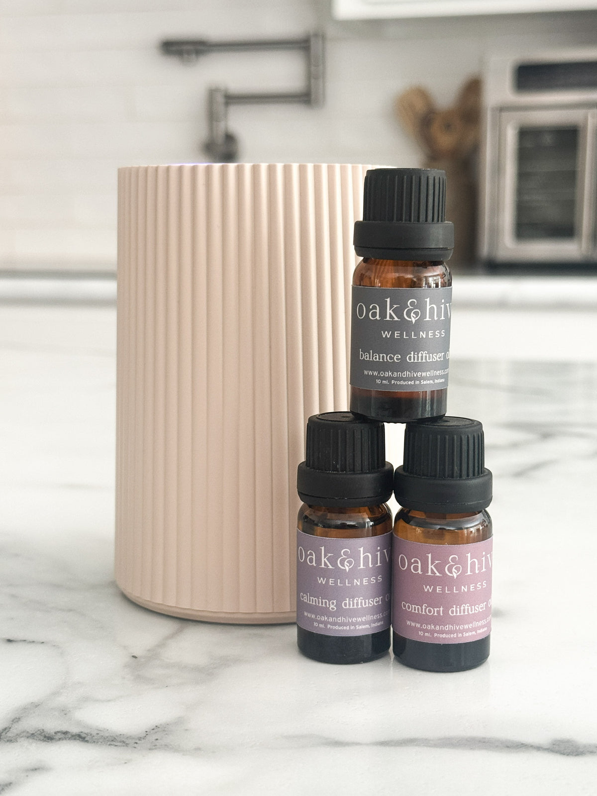 Three amber glass essential oil bottles with black caps and lavender labels are arranged in front of a tall ribbed blush pink cylindrical diffuser on a white marble countertop in a kitchen setting.