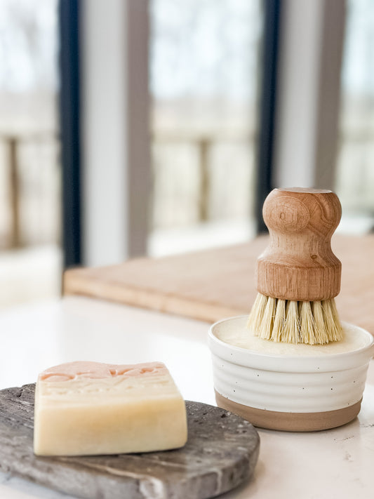 Bar of soap on a stone soap dish with a wooden-handled brush in a white container on a marble surface.