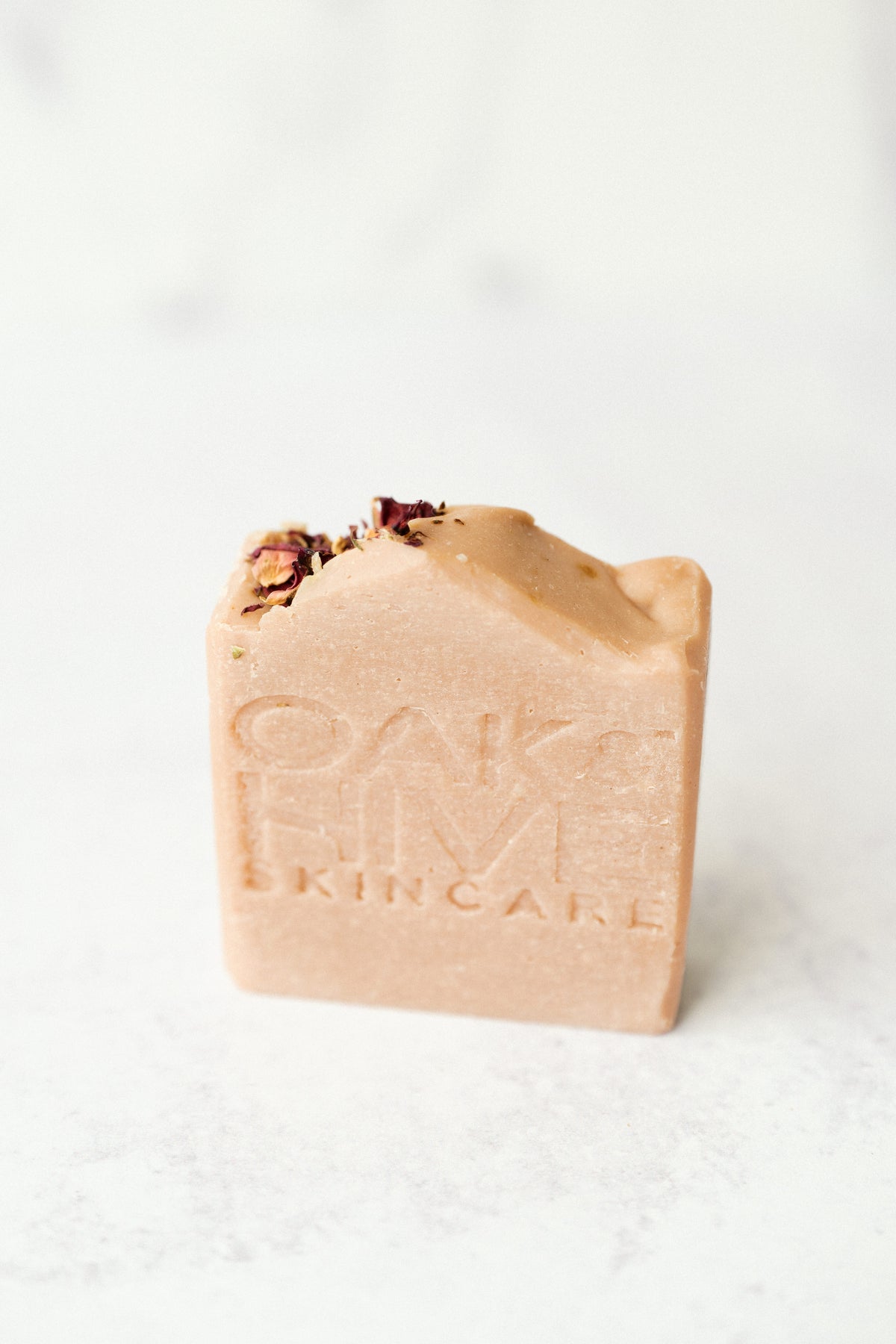 A pink bar of soap with red rose petals on top with a white background. 