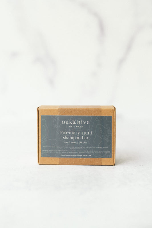 A shampoo bar packaged in a brown paper wrapping with a label that has the brand name 'oak & hive' and the product name 'rosemary mint shampoo bar'. The label is predominantly green and black with text and a small graphic icon.