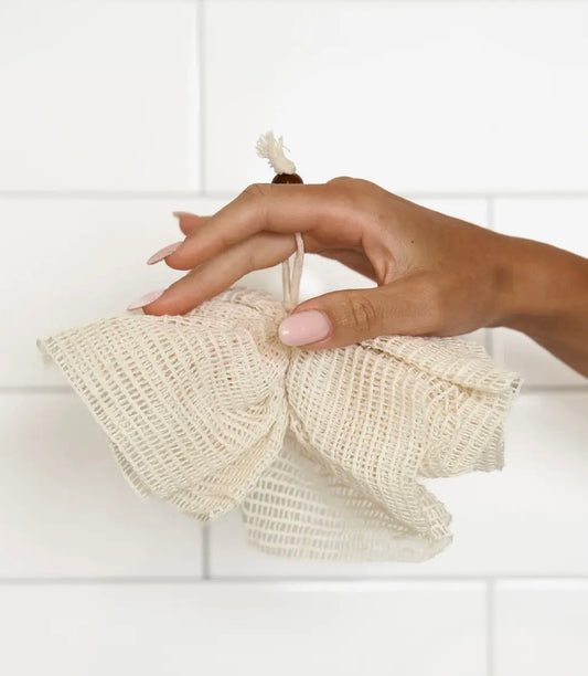 Hand holding a beige reusable sisal shower pouf against a white tiled wall.