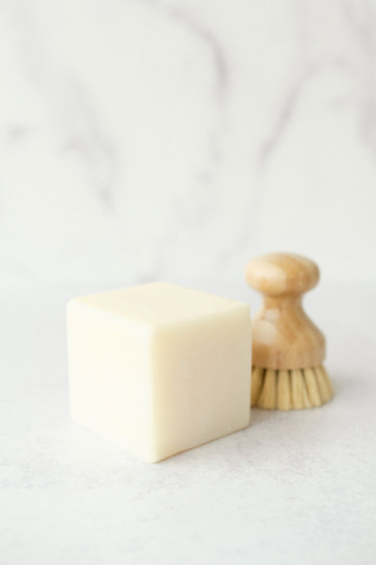 A solid dish soap bar next to a wooden dish brush on a marble surface.