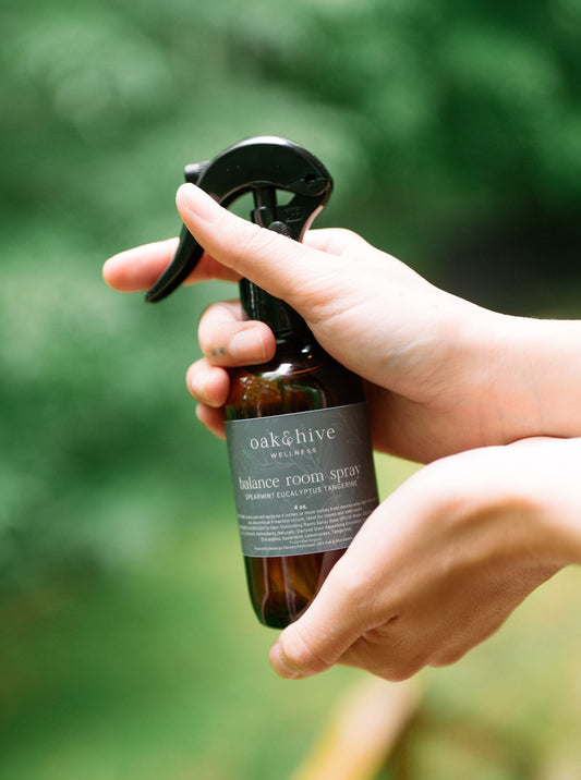 Hand holding a bottle of 'oak&hive' balance room spray with a blurred green background